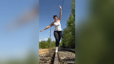 JaneHope — JaneHope balances on a railroad track wearing a white tank top and black leggings under a clear blue sky.