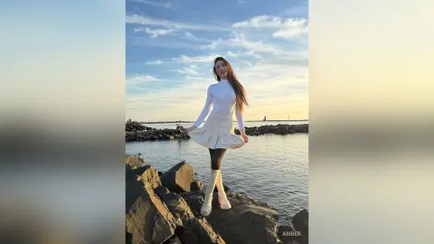 Smiling woman in a white long-sleeve top and skirt standing on rocky coast under a bright blue sky.