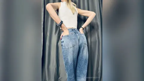 Blonde woman in a white tank top and blue denim jeans standing against a grey curtain backdrop.