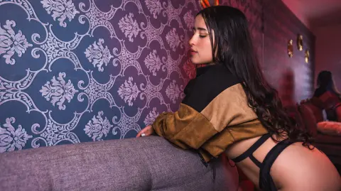Young woman in a brown crop top leaning against a patterned blue wall in moody, warm lighting.