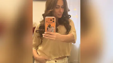 Smiling woman in a beige knit sweater taking a mirror selfie in a cozy indoor setting.