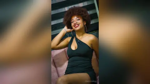 Smiling young woman with afro hair wearing a black halter dress in a cozy indoor setting.