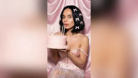 Woman in pink satin corset holding a pink birthday cake against a soft pink satin backdrop.