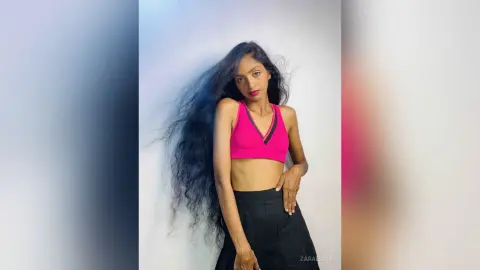 Young woman with long wavy hair wearing a pink crop top and black skirt posing against a white background.