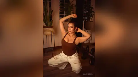 Young woman in a brown bodysuit and ripped white jeans posing in a warm, moody indoor plant setting.