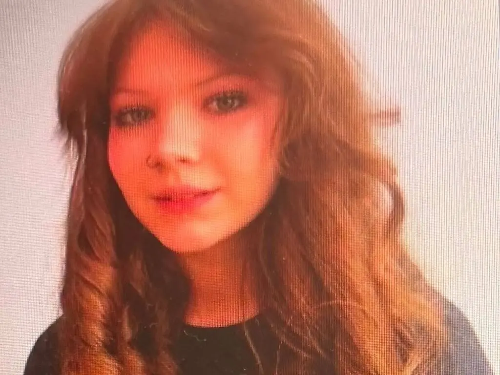 Close-up portrait of a young woman with wavy brown hair wearing a black top against a plain white background.