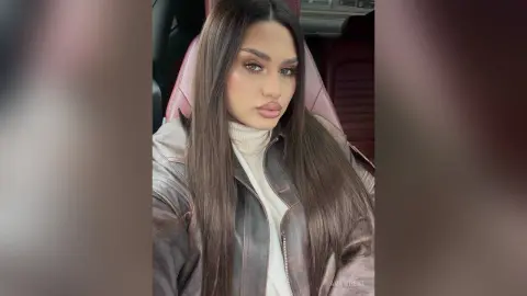 Young woman with long brown hair wearing a white turtleneck and leather jacket sitting in a car.