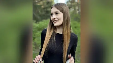 Smiling young woman in a black long-sleeve shirt posing in a lush green forest setting.