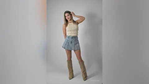 Young woman in a white knit halter top and denim mini skirt posing against a grey studio background.
