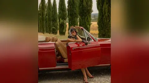 Elegant woman in black chic outfit and sunglasses sitting in a vintage red convertible car in a sunny countryside.