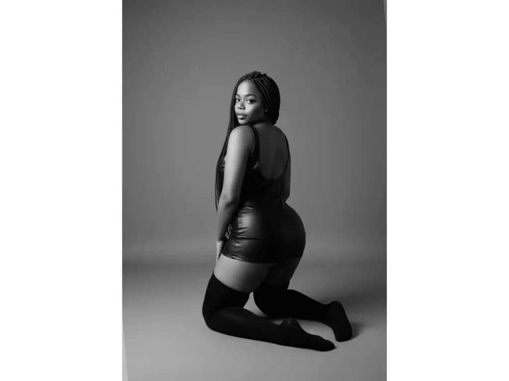 Elegant woman in a black leather mini dress and thigh-high boots posing in a minimalist studio setting.