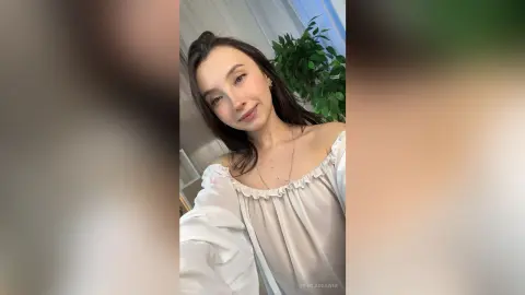 Smiling young woman in a white off-the-shoulder blouse posing in a bright, airy indoor setting.