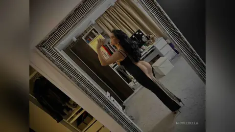 NicoleBella poses in a black dress that exposes her bare buttocks while she stands in a room with a white desk and shelving.