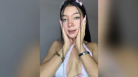 AleDilucas has long dark hair and wears a white halter top with pink trim, posing with her hands against her face.