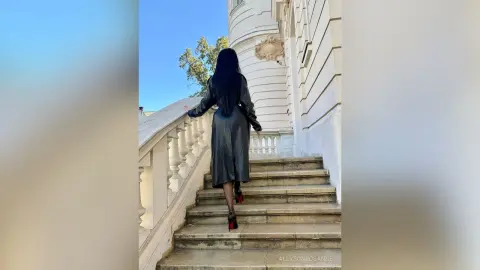 Elegant woman in a long black leather coat walking up white stone stairs of a luxury classical building.