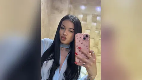 AriaVex winks and pouts her lips while taking a mirror selfie in a light blue shirt with long black hair.