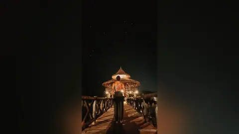 Fit woman in black trousers standing on a wooden pier under a starry night sky at a tropical resort.