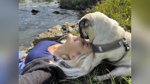 Happy blonde woman in a blue top relaxing outdoors by a river with her playful pug dog.