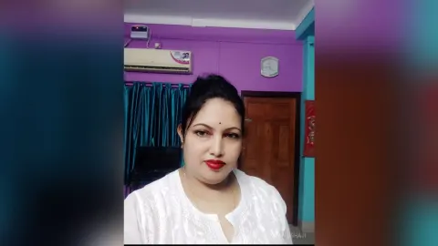 ManishaJi — ManishaJi looks at the camera wearing a white blouse in a room with purple walls.