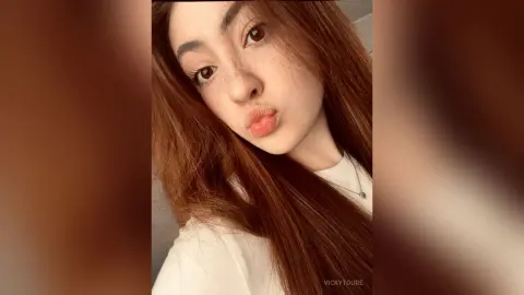 VickyToure pouts her lips and looks at the camera with long brown hair and a white t-shirt.