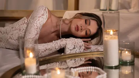 Elegant woman in white lace gown lying gracefully beside glowing candles in a romantic, moody setting.