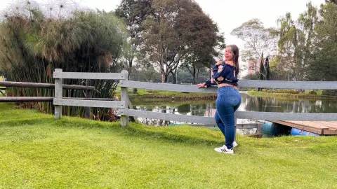 Smiling woman in a black floral top and blue jeans standing by a wooden fence near a peaceful lake.