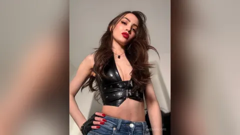 AlienaMoore — AlienaMoore poses in a black leather halter top and blue denim jeans with long brown hair and red lipstick.