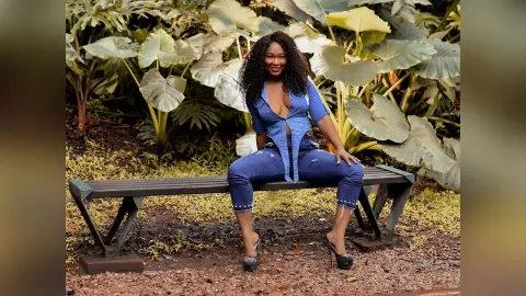 Smiling woman in a blue button-down shirt and denim jeans sitting on a garden bench amidst lush tropical plants.