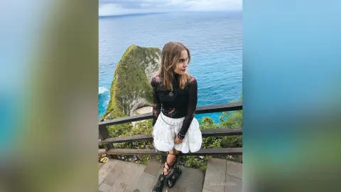 Young woman in a black long-sleeve top and white skirt standing on a scenic coastal cliff overlooking the ocean.