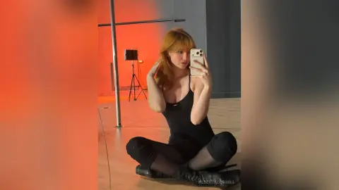 AlbertaCorrell sits cross-legged on the floor in a black tank top and leggings, taking a mirror selfie in a studio with orange lighting.