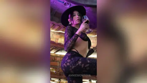 Glamorous woman in a black lace outfit and wide-brimmed hat sipping wine in a moody, dimly lit lounge.