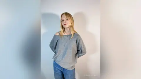 Blonde woman in a grey oversized sweater and blue jeans posing against a white wall with a soft, natural mood.