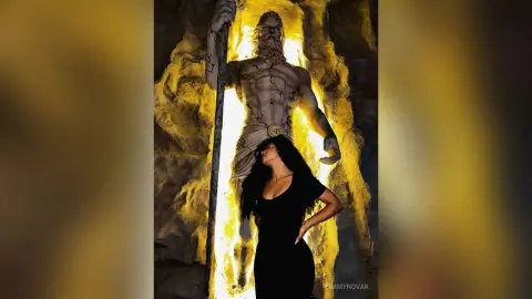 Woman in a black dress standing before a glowing golden statue of a muscular Greek god in a dark cave.