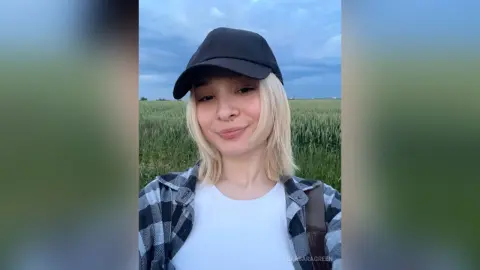 BarbaraGreen wearing a black baseball cap and a black and white plaid shirt, standing in a green field under a cloudy sky.