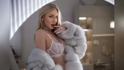 BellaBronze poses in a white lace bra and a thick, fluffy grey fur coat, showing off her blonde hair and light skin.