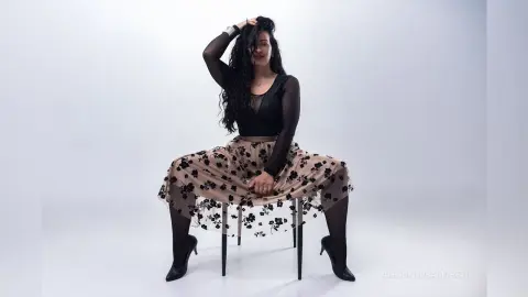 Woman in a black bodysuit and floral midi skirt posing elegantly on a chair against a white studio background.