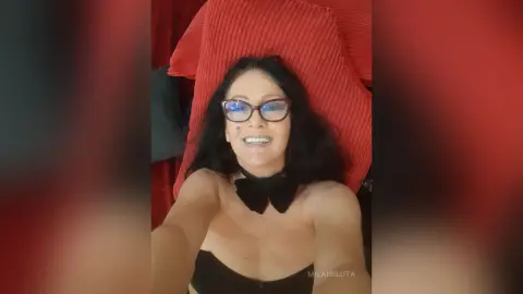 Smiling woman with dark hair and glasses wearing a black strapless top and bow accessory while relaxing on red pillows.