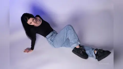 Young woman in black long-sleeve shirt and light wash denim jeans posing against a white studio background.
