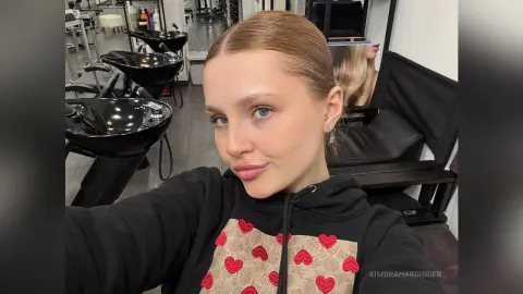 Smiling young woman in a black hoodie posing for a selfie in a modern hair salon setting.