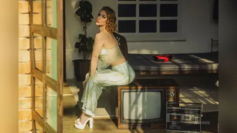 Elegant woman in a light green lace outfit sitting in a sunlit vintage room with a retro television.