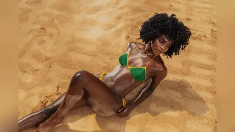 Smiling Black woman in a green crochet bikini relaxing on a sunny sandy beach.