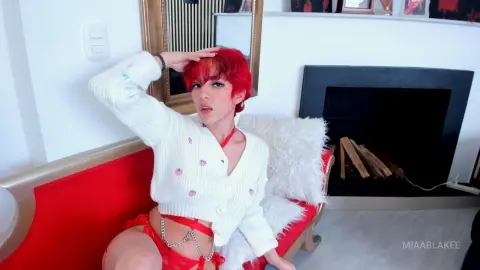 Red-haired woman in a white cardigan and red lingerie relaxing on a red sofa in a bright living room.