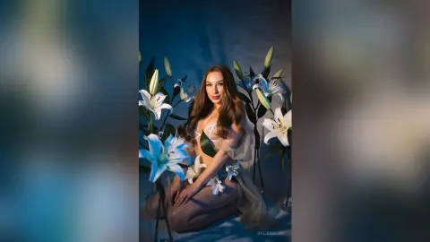 Beautiful woman in a sheer floral outfit posing amidst white lilies in a dreamy, ethereal blue setting.