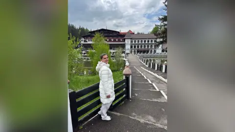 AnishaBee stands outdoors on a paved walkway next to a black fence, wearing a white puffer jacket and white pants with her hair pulled back.