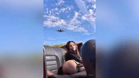 Young woman in a black top relaxing in a car under a bright blue sky with an airplane flying overhead.