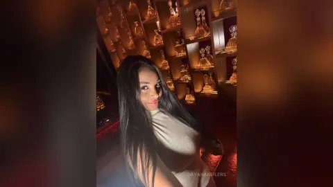 Smiling young woman in a white ribbed top posing in a warm, dimly lit restaurant with glowing decor.