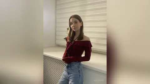 Young woman in a burgundy off-the-shoulder top and blue jeans posing in a bright, minimalist indoor setting.