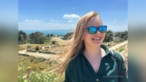 Smiling blonde woman in a dark green shirt and blue sunglasses enjoying a sunny day at an ancient coastal ruin.