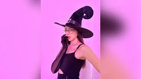 Young woman in witch costume and glasses posing against a pink background for a spooky Halloween mood.