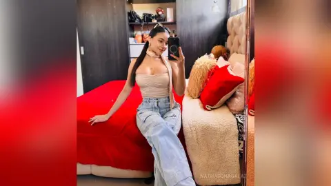 Young woman in a beige halter top and light blue jeans sitting on a red bed in a cozy bedroom setting.